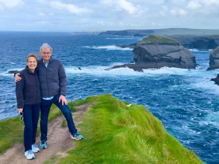 An older couple stands smiling on a grassy cliff overlooking the ocean, with rocky sea stacks and waves in the background under a partly cloudy sky.