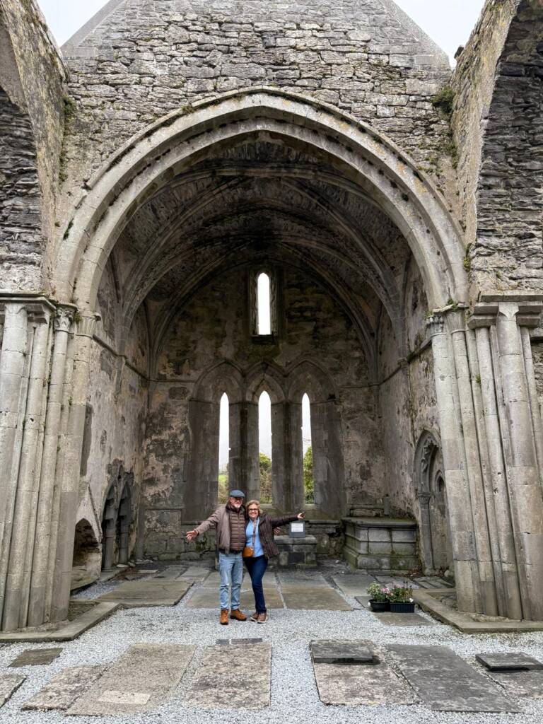 Two people stand smiling with arms outstretched inside the ruins of an old stone church or abbey with tall arches, vaulted ceilings, and narrow windows. Weathered stone walls and monuments surround them.
