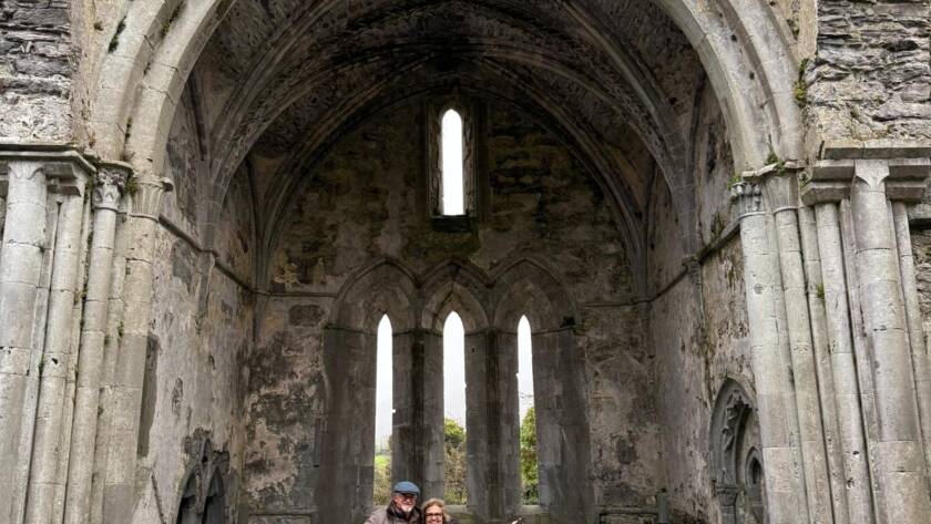 Two people stand smiling with arms outstretched inside the ruins of an old stone church or abbey with tall arches, vaulted ceilings, and narrow windows. Weathered stone walls and monuments surround them.