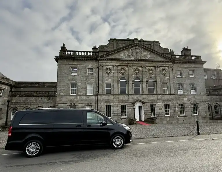 A large, elegant stone building with arched windows and decorative details stands behind a black van parked on the street in front. The sky is cloudy, and sunlight is visible in the background.