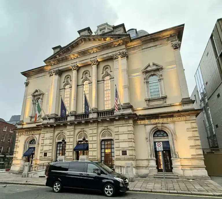 A large, elegant stone building with tall arched windows and columns, illuminated by exterior lights. Flags are displayed above the entrance, and a black van is parked on the street in front.