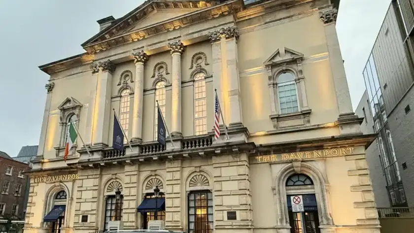 A large, elegant stone building with tall arched windows and columns, illuminated by exterior lights. Flags are displayed above the entrance, and a black van is parked on the street in front.