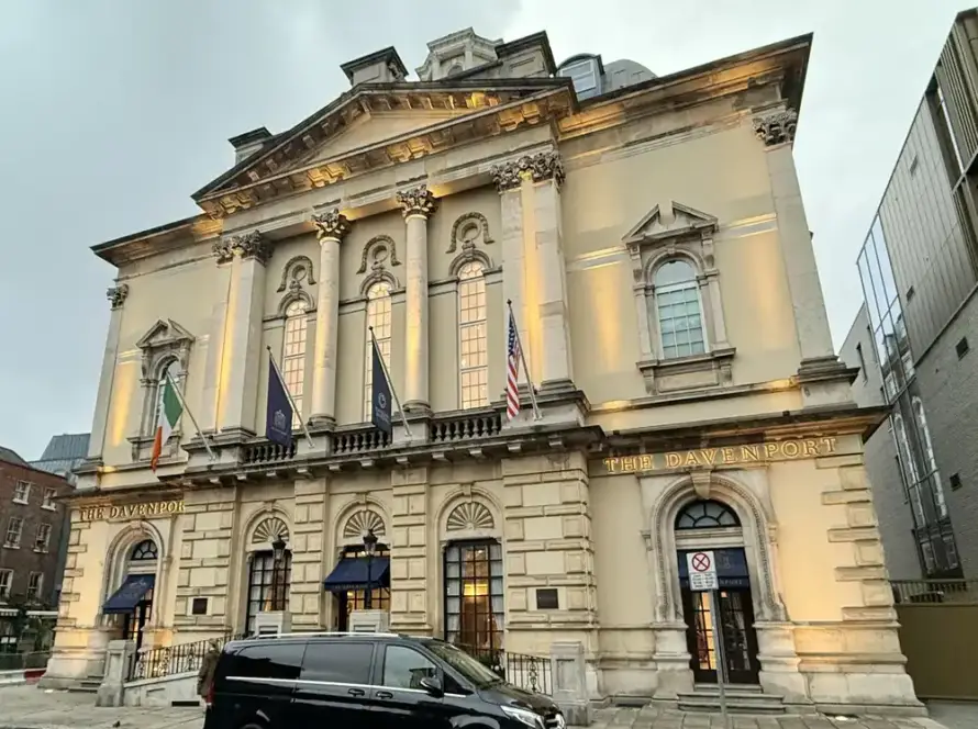 A large, elegant stone building with tall arched windows and columns, illuminated by exterior lights. Flags are displayed above the entrance, and a black van is parked on the street in front.