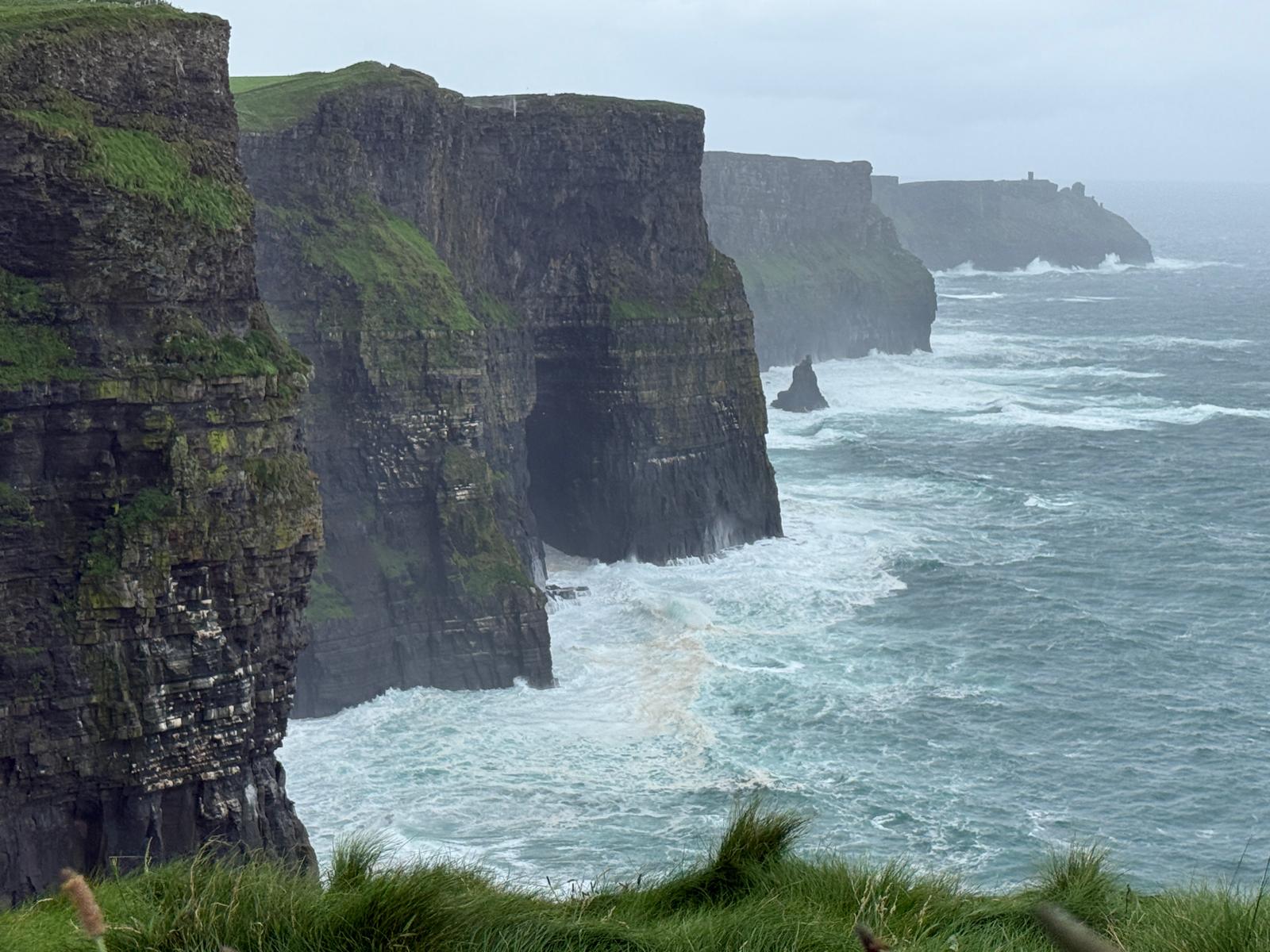 Steep, rugged cliffs rise above crashing ocean waves under a cloudy sky, with green grass covering the Cliffs of Moher. The sea appears choppy, and the distant cliffs fade into the mist.