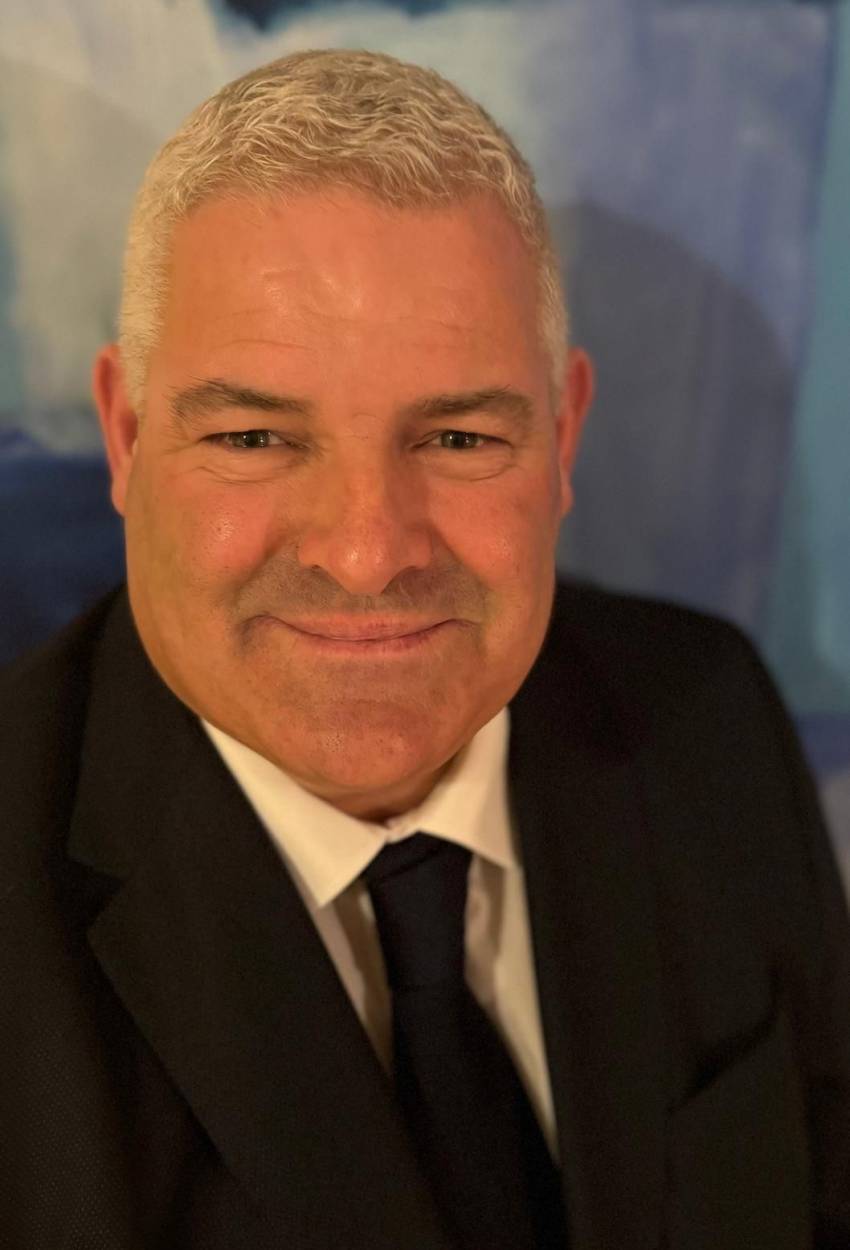 A middle-aged man with short gray hair, wearing a black suit, white shirt, and black tie, smiles at the camera against a blurred blue background.