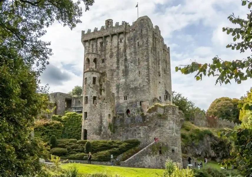 A large stone castle with tall walls and battlements stands on a grassy hill, surrounded by trees and greenery, under a partly cloudy sky. Several people are visible exploring the grounds.